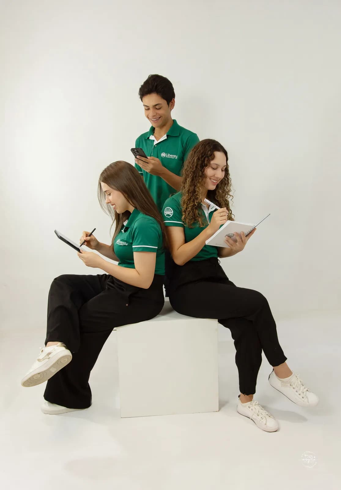 Três integrantes do time Liberty de polo verde, sentadas e em pé, trabalhando com prancheta e celular em ensaio de estúdio